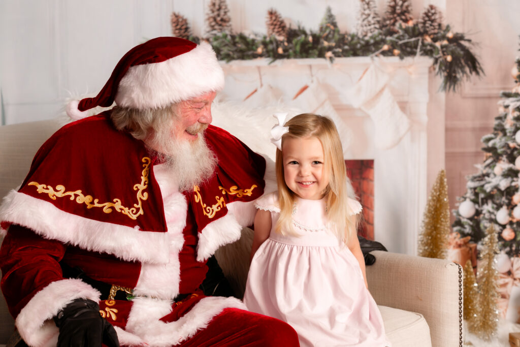 little girl with blonde hair sitting on a sofa next to Santa. Santa is smiling at her and she is smiling at the camera. Columbia, SC Santa Photos