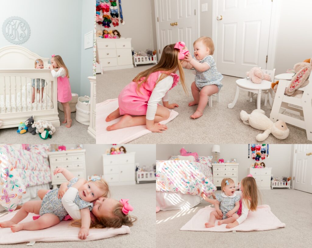 Big sister in a pink dress playing with her baby sister in a bright nursery, helping her with a bow, hugging, and laughing together during an in-home family photo session in Columbia, SC.