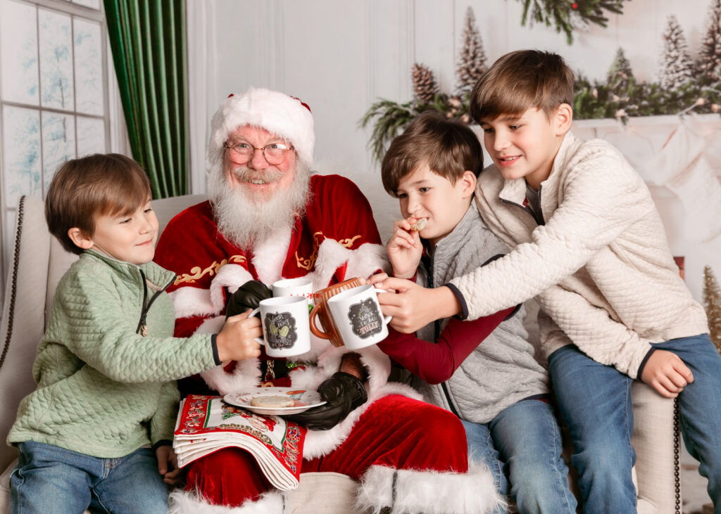Santa photo sessions in Columbia SC – Santa Claus sharing cookies and hot cocoa with three smiling boys during festive holiday portraits. Magical Christmas photos by Emily Ann Photography.