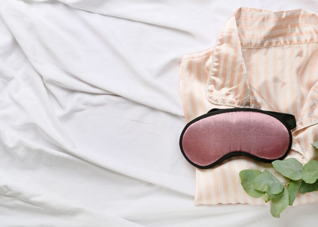 white sheets with a pink and white striped silk pajama shirt folded and a pink silk eye mask laying on top