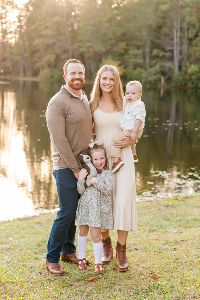 family stands in front of water posing for portraits. baby in moms arms, toddler holding her stuffed animal