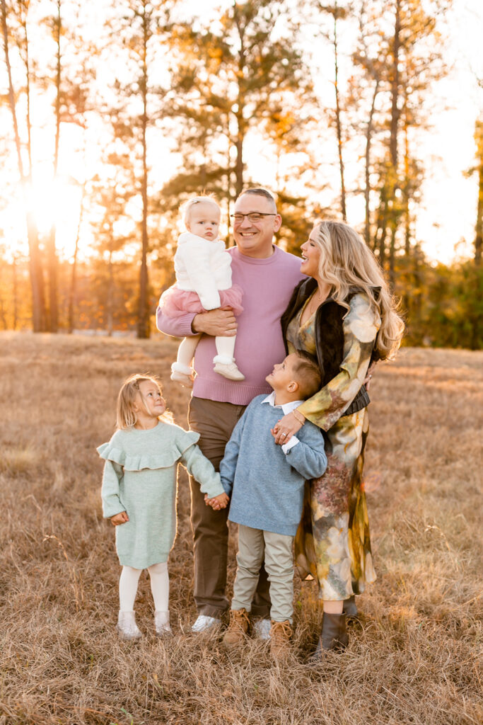 Family portrait in Columbia SC at sunset, parents holding baby with two young children in a field.
